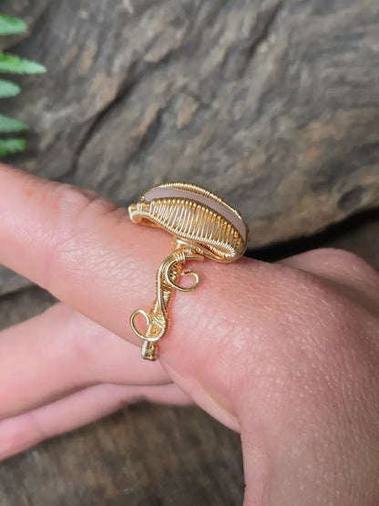A gold copper wrap ring with a rose quartz gemstone, worn on a person's finger against a natural wood and green leaf background.