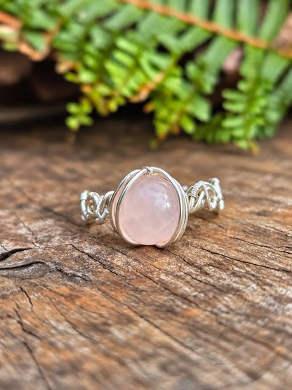 Handmade silver-coloured copper wire wrapped ring on a natural wooden surface with green ferns in the background 