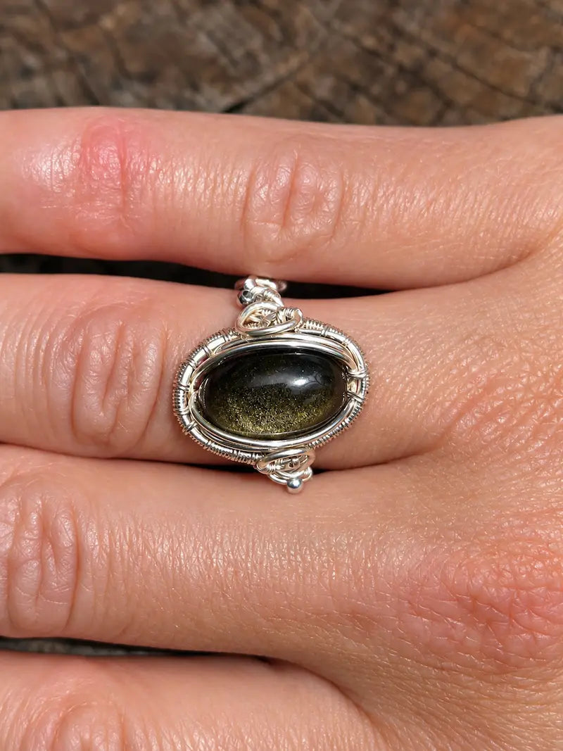 A silver copper ring with an oval dark green stone, shown worn on a person's finger against a textured brown background.