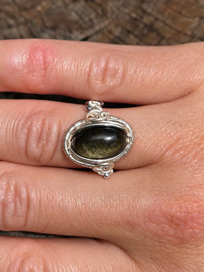 A silver copper ring with an oval dark green stone, shown worn on a person's finger against a textured brown background.
