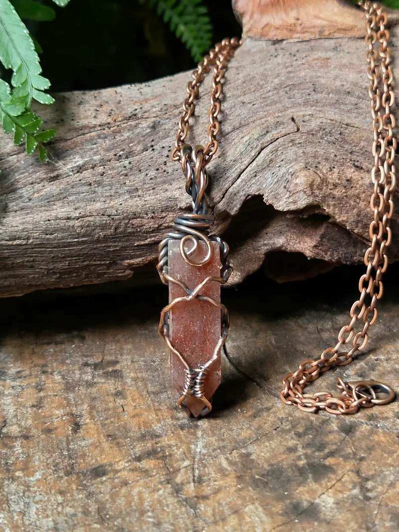 A handmade copper wire-wrapped necklace featuring a polished brownish-red stone pendant, displayed on a rustic wood log with green leaves in the background.