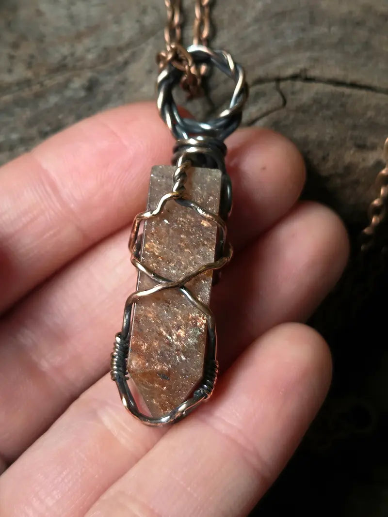 A close-up of a hand holding a handmade wire-wrapped crystal pendant necklace. The pendant features a hexagonal brown crystal wrapped in copper wire, suspended from a twisted copper-toned chain against a dark, rustic wood background.