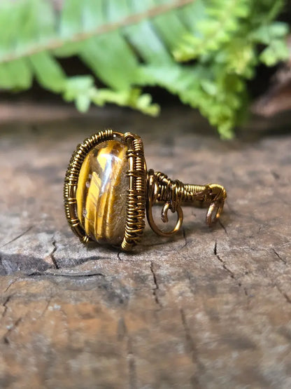 A handmade copper wire-wrapped ring featuring a polished tiger's eye bead, resting on a textured wooden surface with a soft-focus background of green fern leaves.