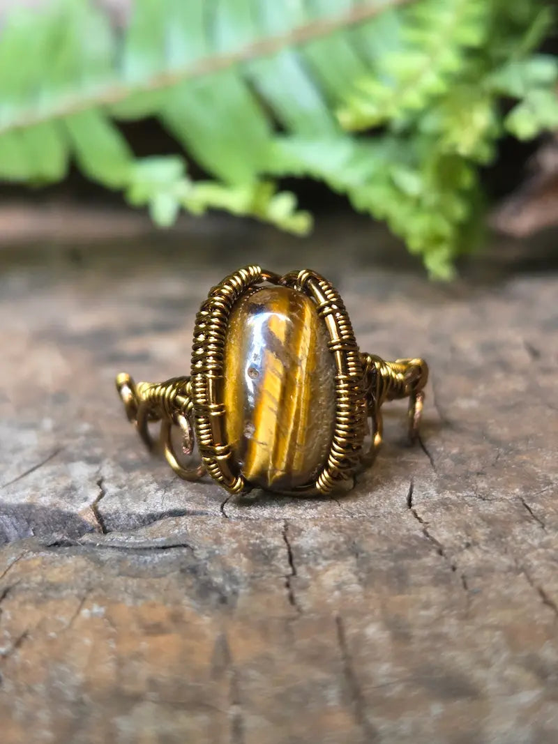 A handmade copper wire-wrapped ring featuring a polished tiger's eye stone, resting on a textured wooden surface with green fern leaves in the background.