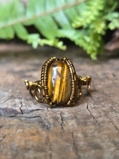 A handmade copper wire-wrapped ring featuring a polished tiger's eye stone, resting on a textured wooden surface with green fern leaves in the background.