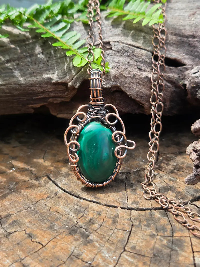 Copper pendant necklace with a green stone, resting on a natural wooden surface with green ferns in the background 