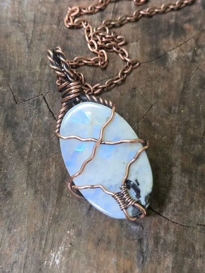 A handmade oval pendant necklace featuring a light blue iridescent moonstone wrapped in copper wire, resting on a rustic dark wood surface.