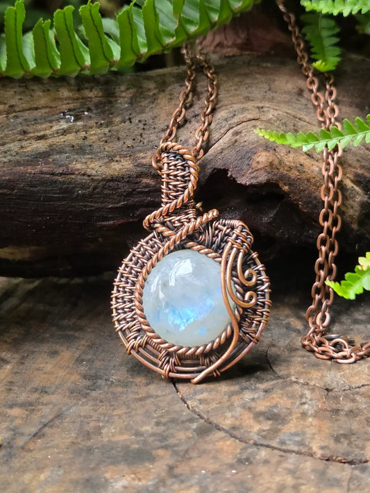 Copper-wrapped necklace with a blue  moonstone pendant on a wooden surface with green leaves.