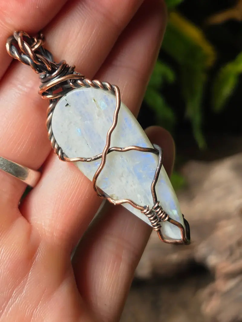 A close-up of a hand holding a teardrop-shaped white moonstone pendant wrapped in copper wire, with a matching copper chain loop at the top. The background is a soft-focus outdoor scene with green foliage and brown rocks.