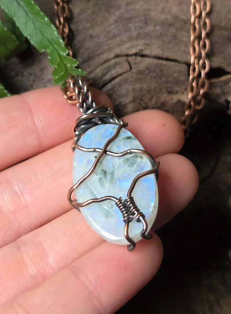 A close-up of a hand holding a oval-shaped iridescent white stone pendant wrapped in dark copper wire, with a copper chain visible against a rustic wood and green leaf background.