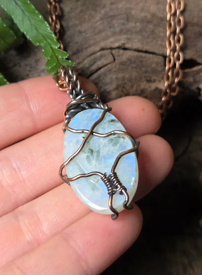 A close-up of a hand holding a oval-shaped iridescent white stone pendant wrapped in dark copper wire, with a copper chain visible against a rustic wood and green leaf background.