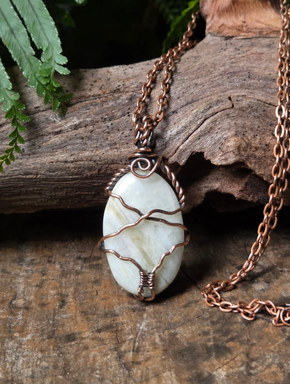 An oval pendant necklace with a pale white stone wrapped in copper wire, resting on a rustic piece of wood with a fern in the background.