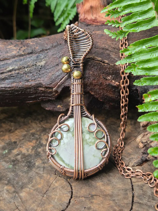 Copper wire-wrapped pendant with a green stone on a wooden surface with ferns in the background