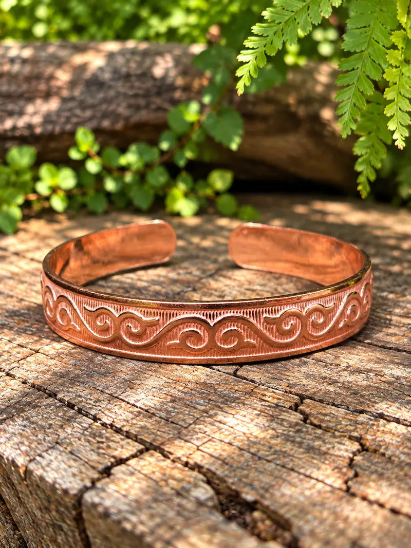 A copper cuff bracelet with engraved scrollwork patterns, resting on a weathered wooden log with green foliage in the background.