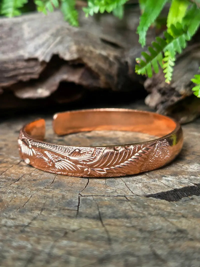 A copper cuff bracelet with a leaf design, resting on a natural wooden surface with green leaves in the background 