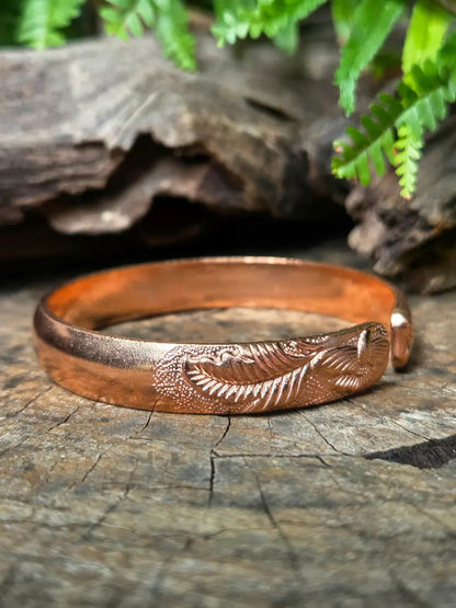 A copper cuff bracelet with an engraved leaf pattern, resting on a weathered wood surface with green ferns in the background.