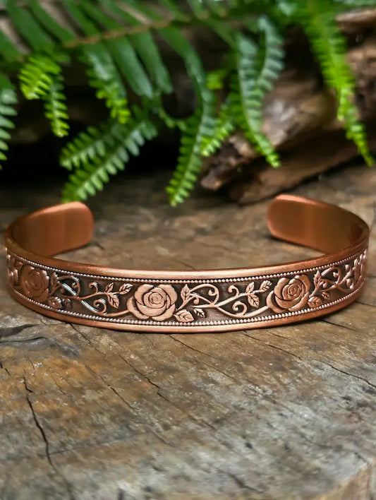 Copper bracelet with floral rose patterns on a natural wooden surface with green ferns in the background 