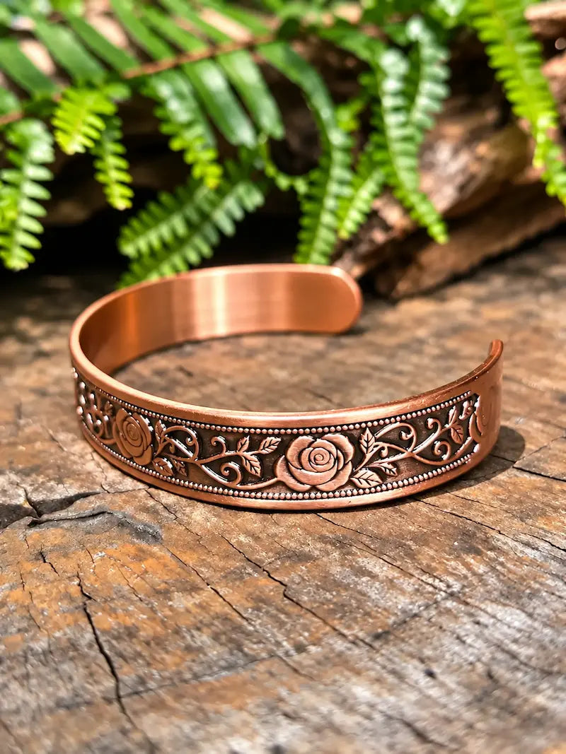 Copper cuff bracelet with a floral rose design, on a natural wooden surface with green ferns in the background.