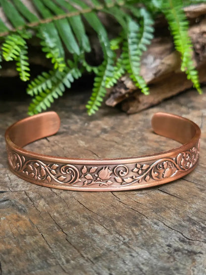 Copper bracelet with a floral rose design on a natural wooden surface with green leaves in the background 