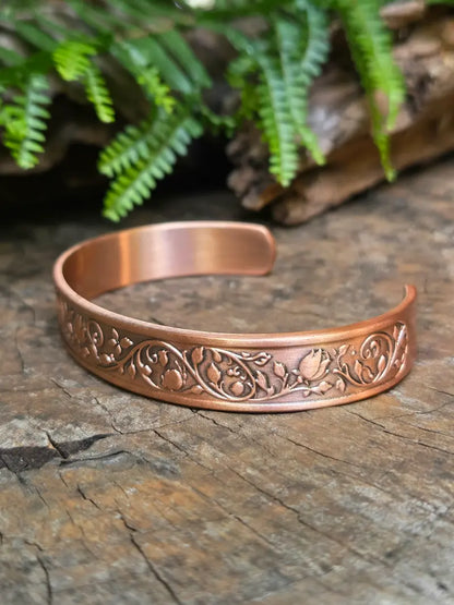 Copper bracelet with a floral rose design on a natural wooden surface with green leaves in the background 