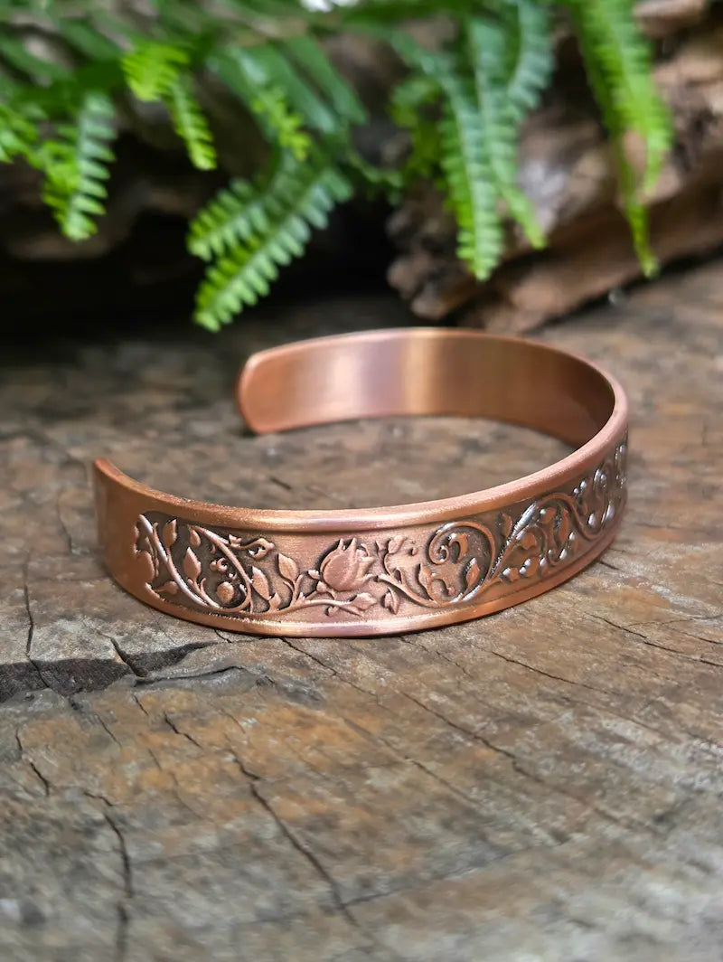Copper bracelet with a floral rose design on a natural wooden surface with green leaves in the background 