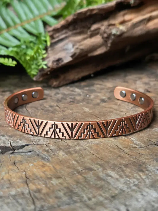 A copper cuff bracelet with engraved geometric patterns, displayed on a rustic wooden surface with blurred green leaves and wood in the background.