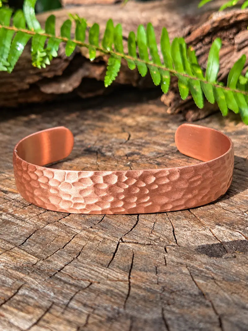 A copper bracelet with a hammered design, resting on a natural wooden surface with green ferns in the background 