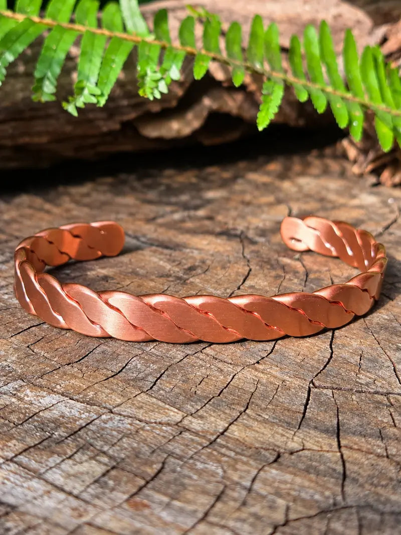 A twisted copper bracelet resting on a natural wooden surface with green ferns in the background 