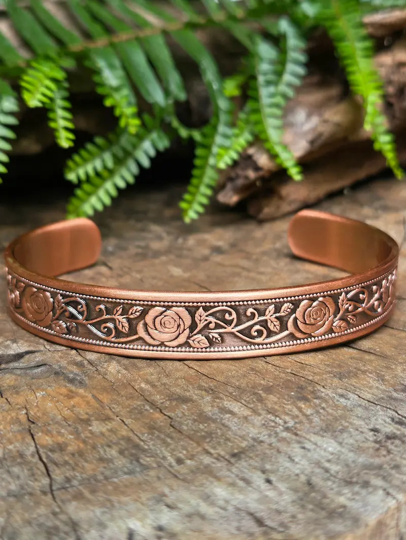 Copper bracelet with a elegant floral rose design on a natural wooden surface with green leaves in the background 