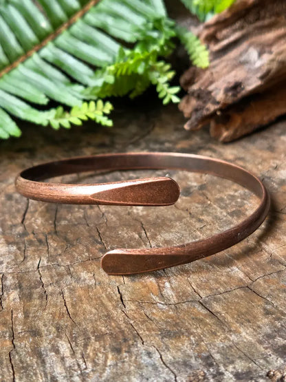 A copper open cuff bracelet resting on a rustic wood surface with green fern leaves and a piece of driftwood in the background.