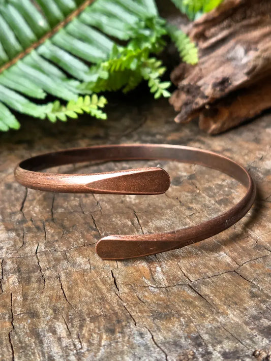 A copper open cuff bracelet resting on a rustic wood surface with green fern leaves and a piece of driftwood in the background.