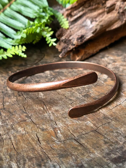 A copper open cuff bracelet resting on a textured wooden surface with green fern leaves and driftwood in the background.