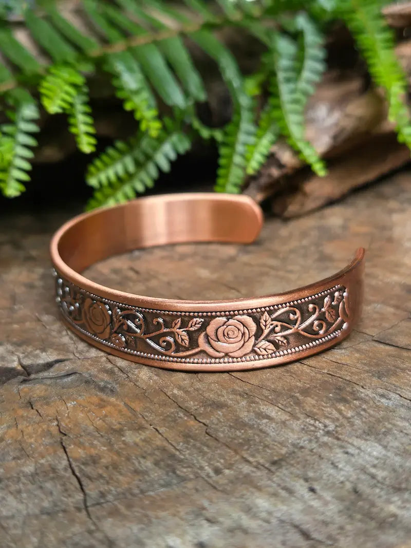 Copper bracelet with a elegant floral rose design on a natural wooden surface with green leaves in the background 