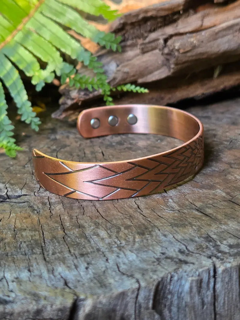 A copper bracelet with a sunburst geometric design, resting on a natural wooden surface with green ferns in the background.