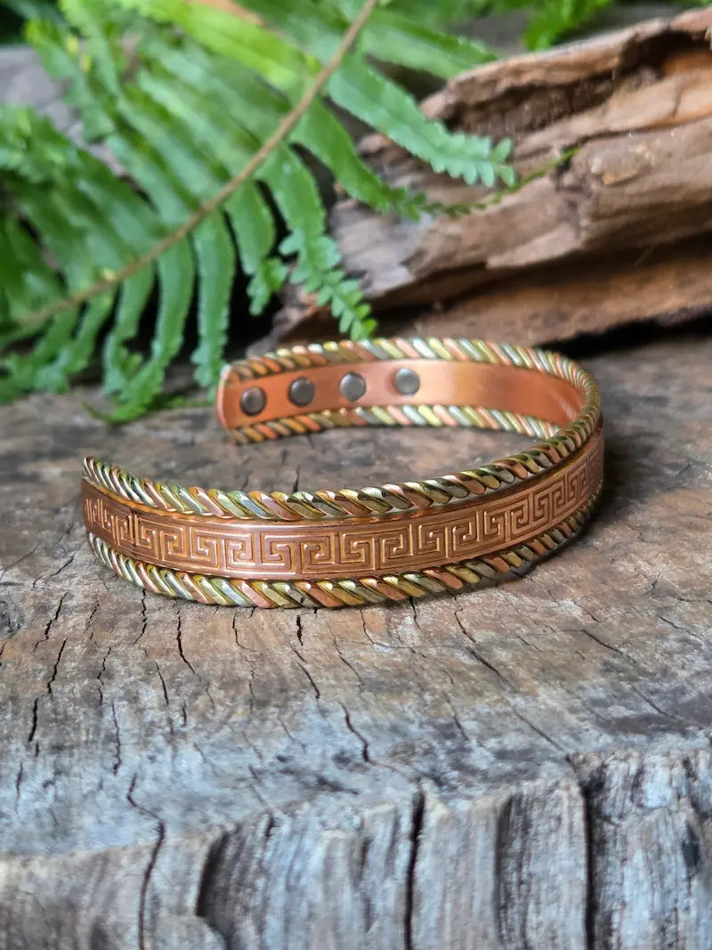 A copper bracelet with a Tricolour greek key design,  resting on a natural wooden surface with green ferns in the background.