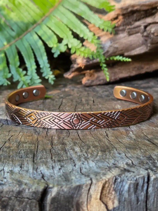 A copper bracelet with a geometric design, resting on a natural wooden surface with green ferns in the background 
