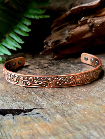 Copper bracelet with a floral heart design on a natural wooden surface with green ferns in the background 