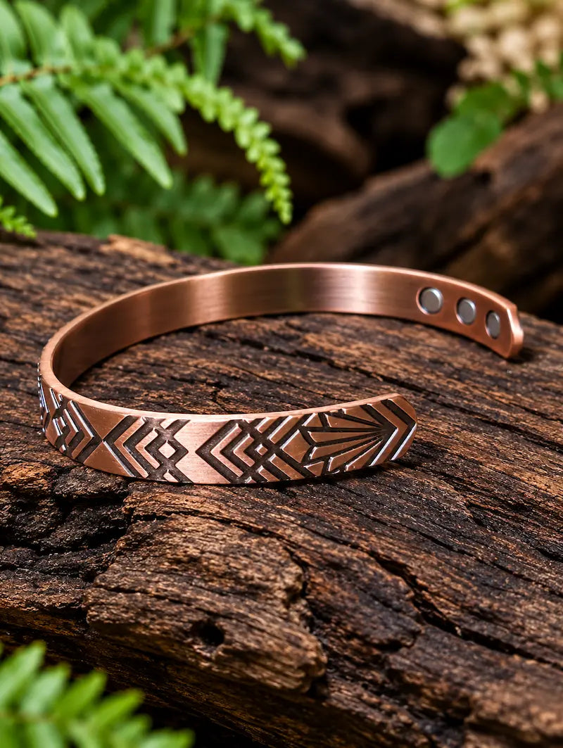 Copper Bracelet with a geometric pattern resting on a natural wooden surface with green ferns in the background 