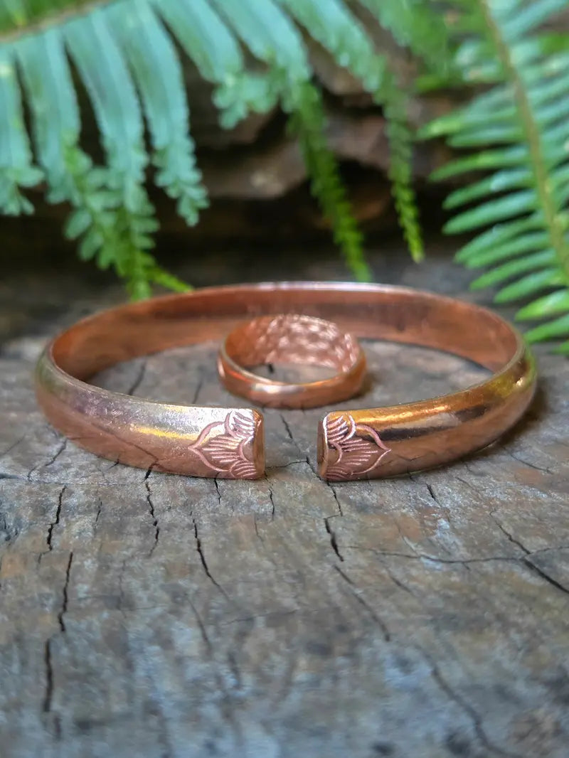 A copper open cuff bracelet and matching ring with textured and engraved floral details, displayed on a weathered wooden surface with green ferns in the background.
