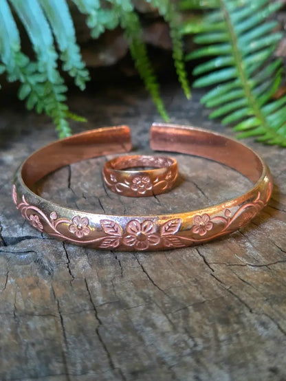 A copper open cuff bracelet with embossed floral patterns, displayed on a weathered wood surface with a matching ring in front and green fern leaves in the background.