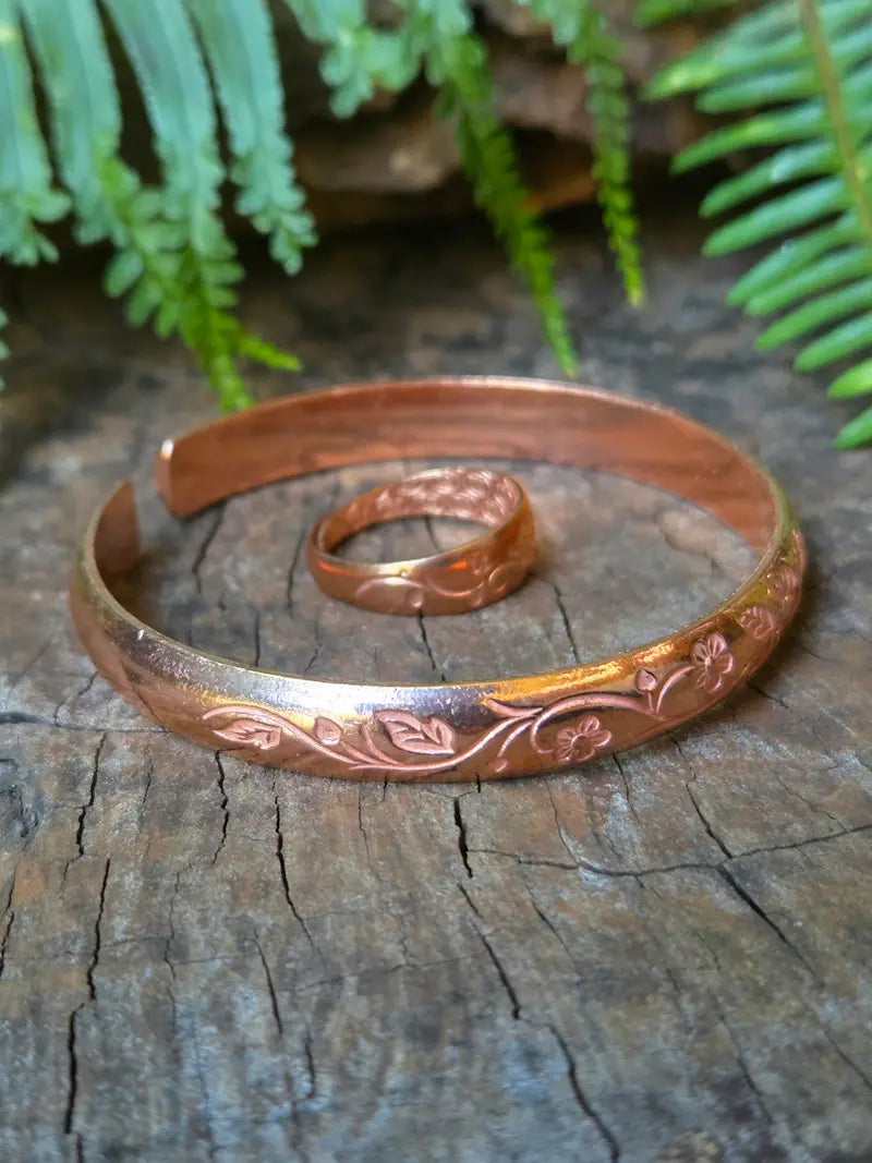 A copper engraved cuff bracelet and matching ring displayed on a weathered wooden surface with green fern leaves in the background.