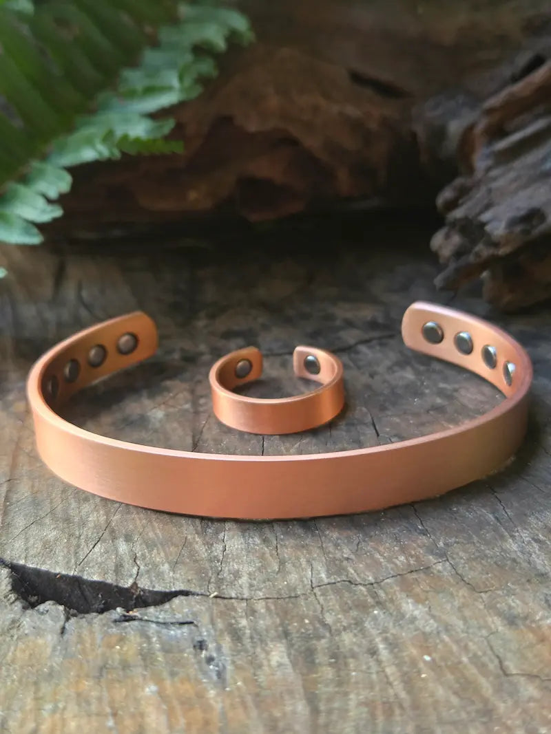 Copper cuff bracelet and ring displayed on a rustic wooden surface with a fern leaf in the background.