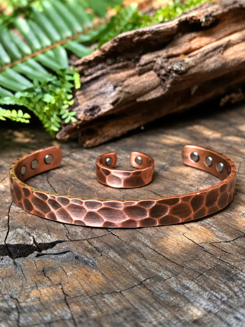 A hammered copper cuff bracelet and matching ring displayed on a rustic wooden surface with green leaves and driftwood in the background.