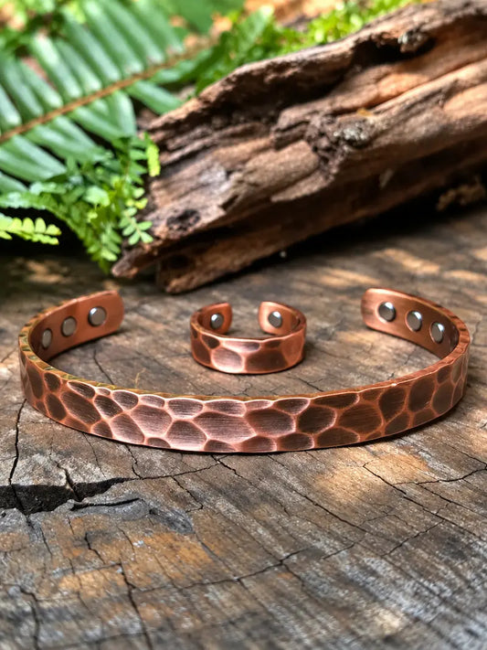 A hammered copper cuff bracelet and matching ring displayed on a rustic wooden surface with green leaves and driftwood in the background.