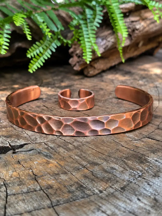 A hammered copper cuff bracelet and matching ring displayed on a rustic wood surface with green fern leaves in the background.