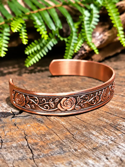 A copper-toned open cuff bracelet with embossed floral and scrollwork details, resting on a rustic wooden surface with green ferns in the background.