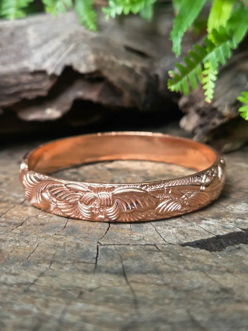 A copper bracelet with an engraved floral pattern, resting on a rustic wood log with green fern leaves in the background.