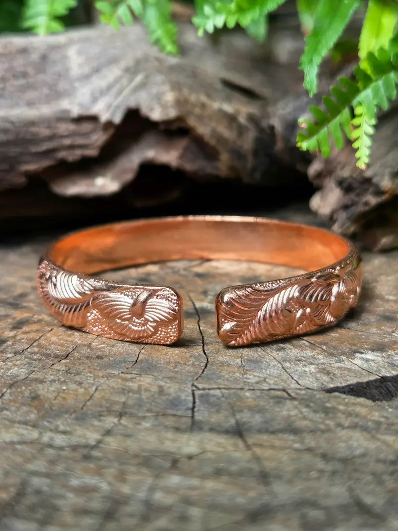 A copper cuff bracelet with engraved feather designs, resting on a textured wooden surface with a blurred natural background of wood and green leaves.