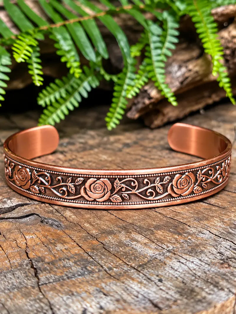 Copper cuff bracelet with a floral rose design, on a natural wooden surface with green ferns in the background.