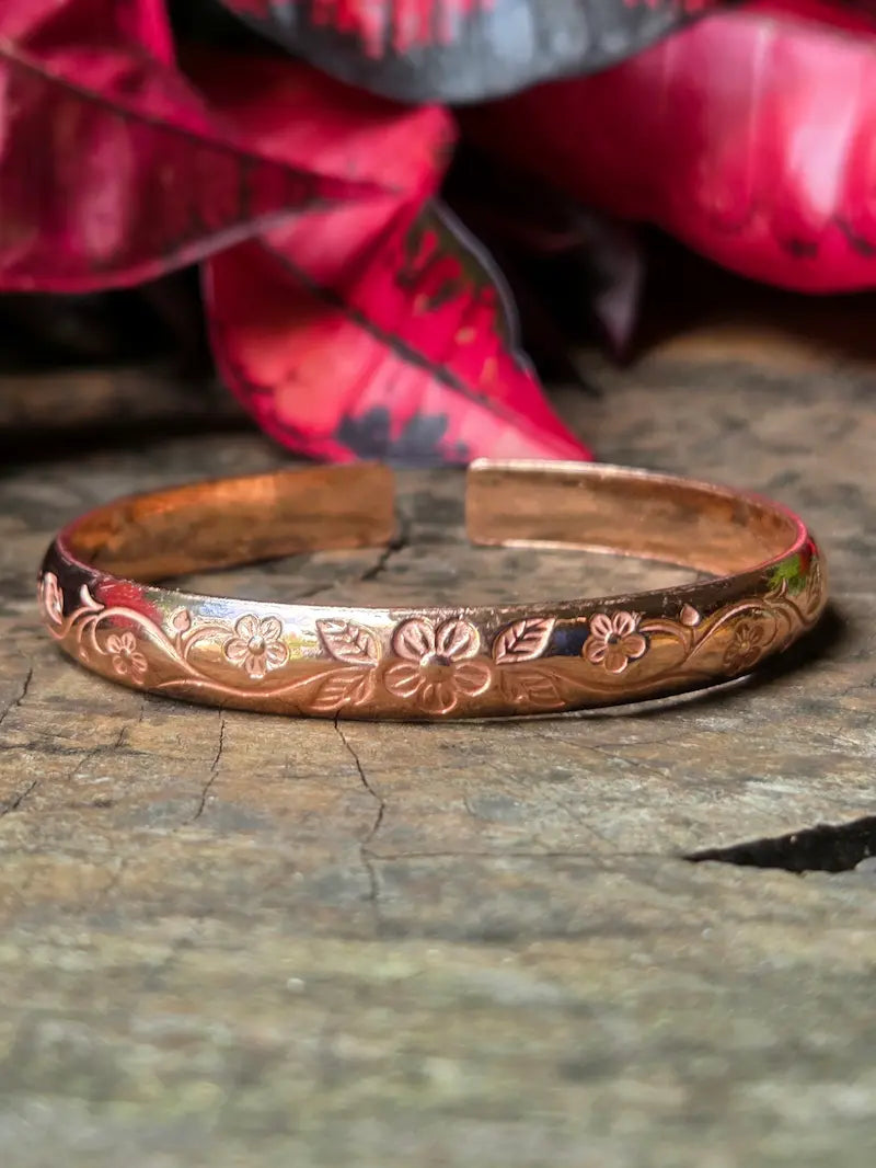 Copper bracelet with floral patterns on a wooden surface with red leaves in the background 